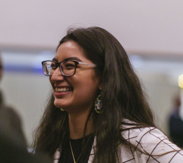 Denisse - young, Latina woman in glasses and blazer - smiling in crowd