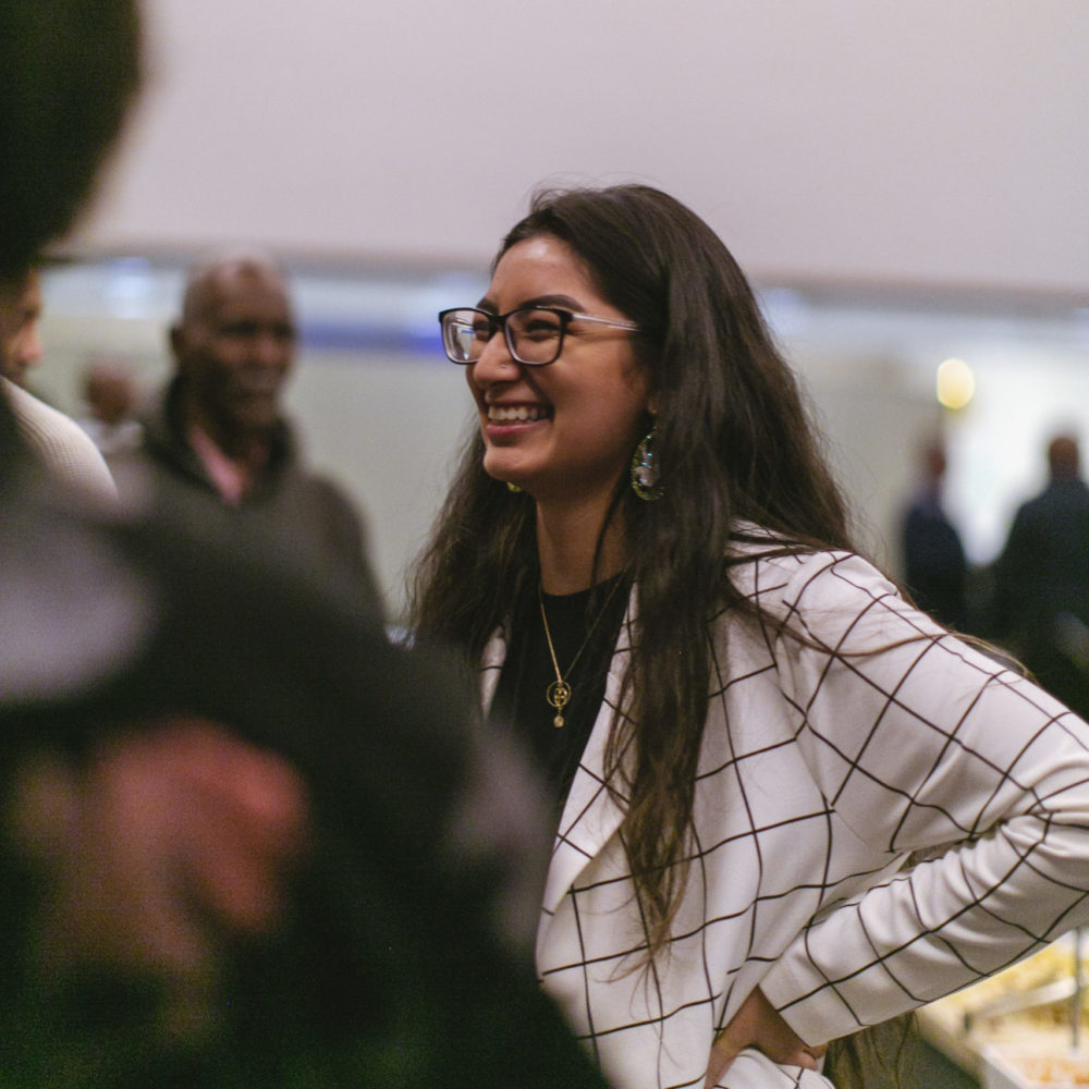 Denisse - young, Latina woman in glasses and blazer - smiling in crowd